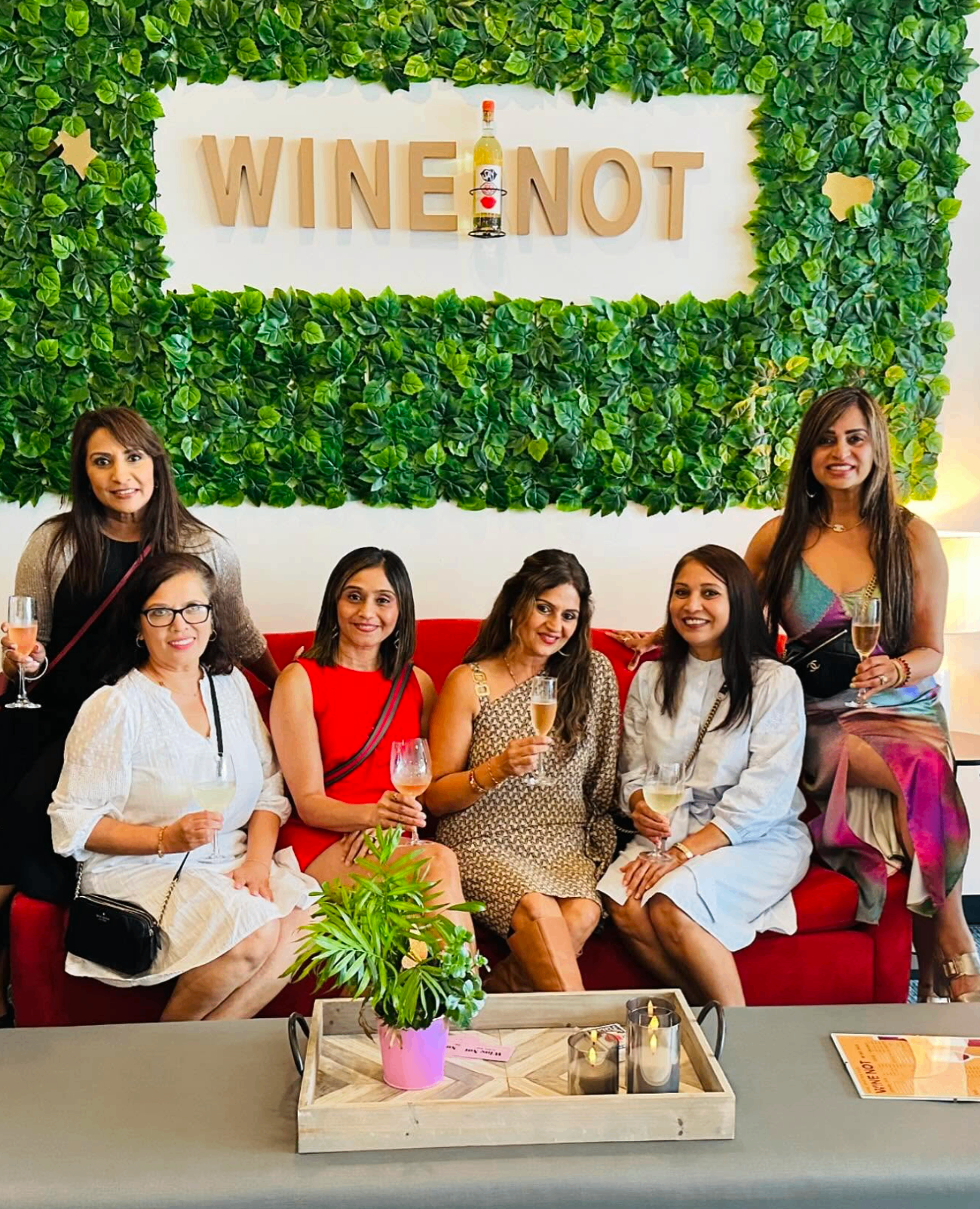 Group of women toasting on the red couch under the WINE NOT greenery wall