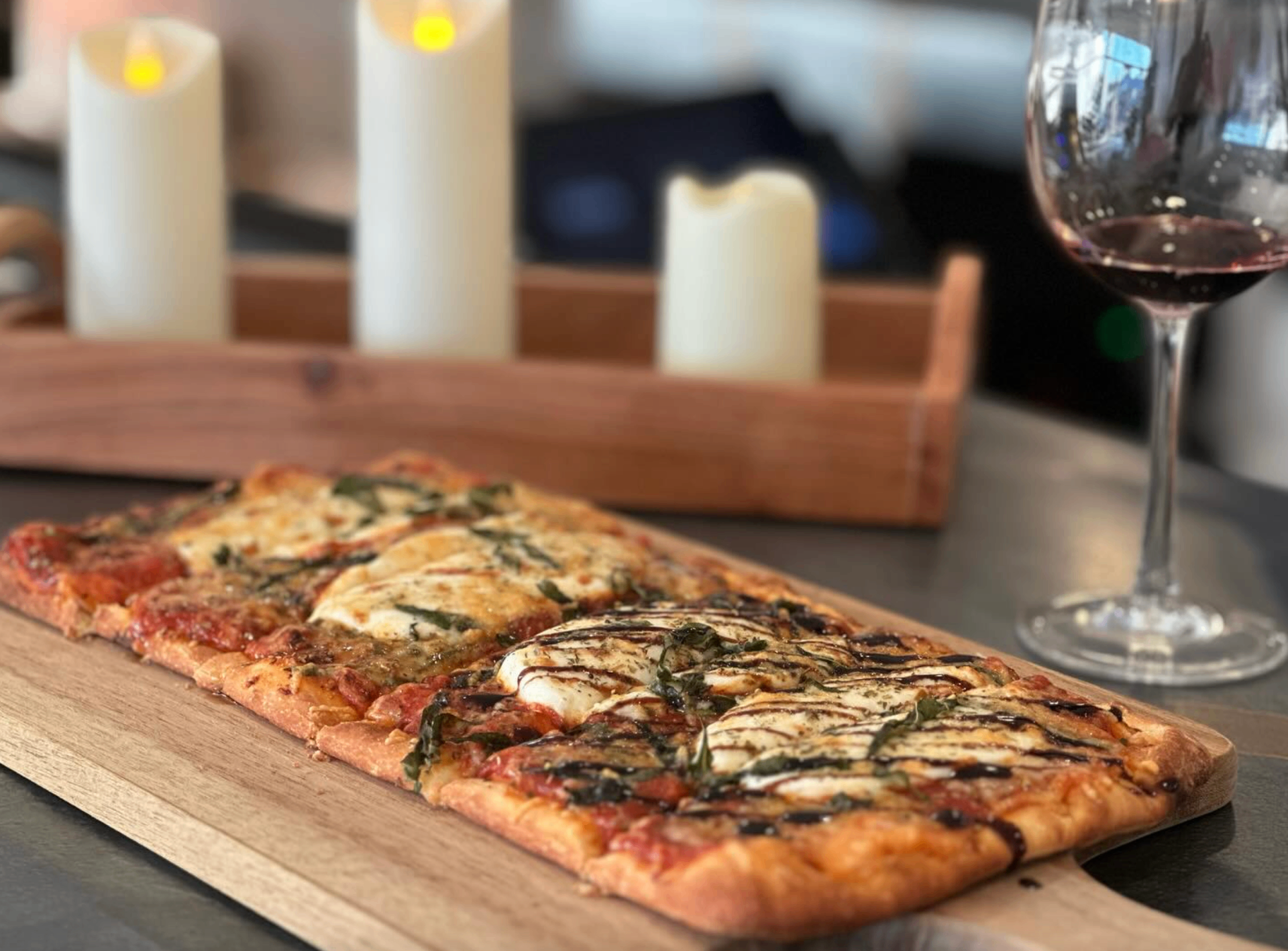 Margherita flatbread with balsamic drizzle on a wooden board next to a glass of red wine and candles
