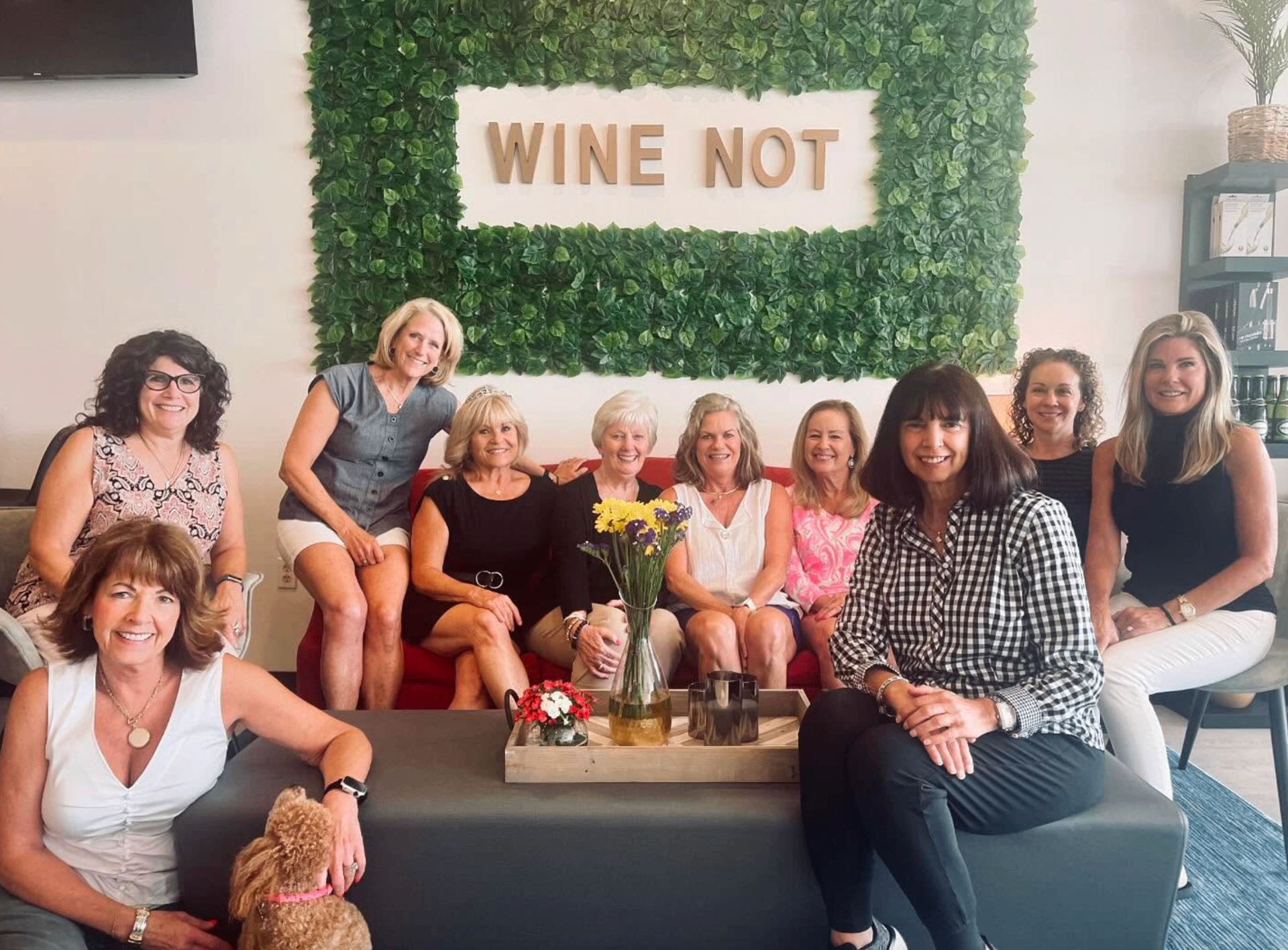 Group of women smiling on the lounge in front of the Wine Not greenery wall