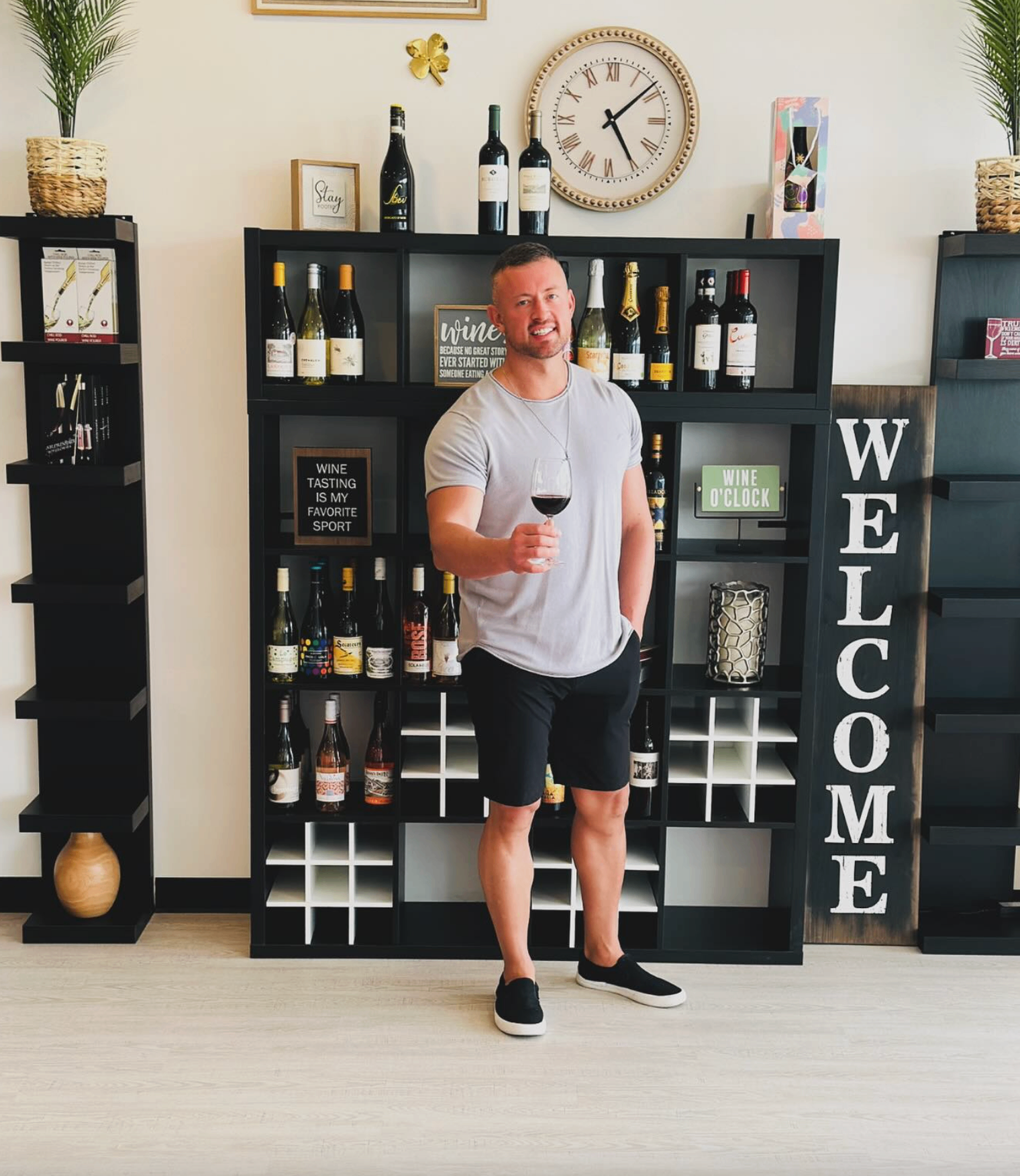 Man holding a glass of red wine in front of the Wine Not bottle shelves and Welcome sign