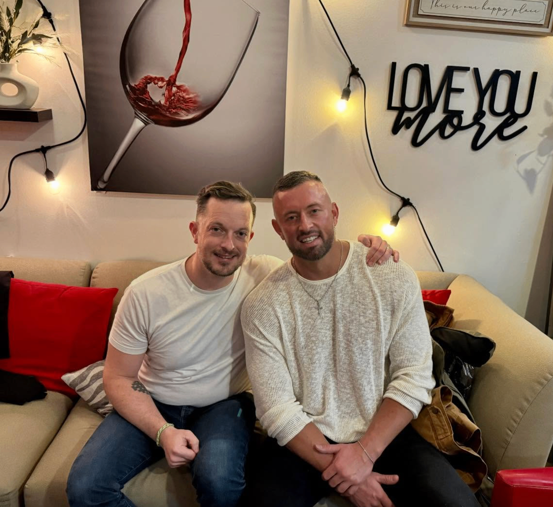 Two friends on the couch under Love You More sign