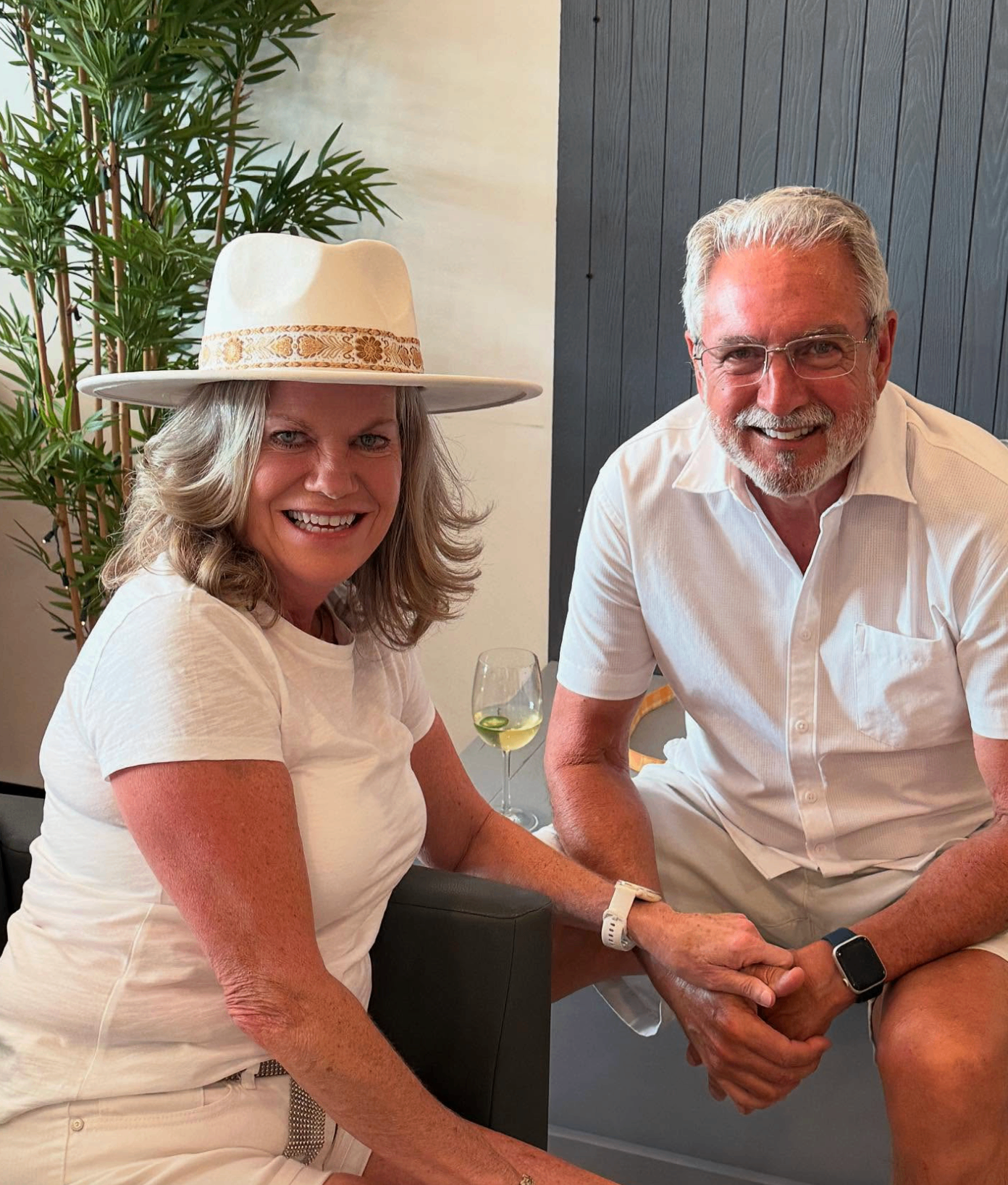 Couple in white smiling with a glass of white wine, woman in a wide-brim hat