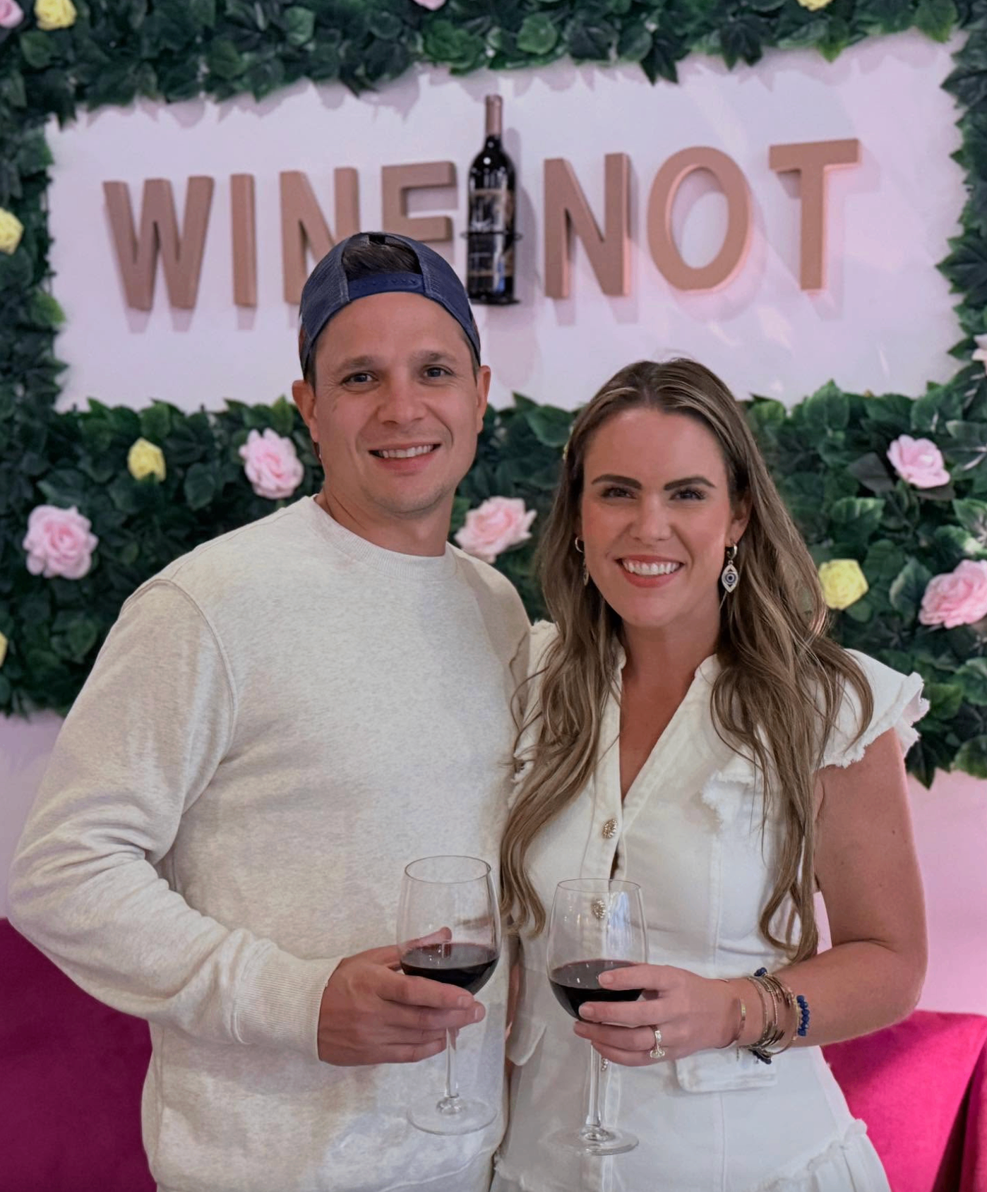 Couple in white outfits toasting with red wine in front of the Wine Not floral wall