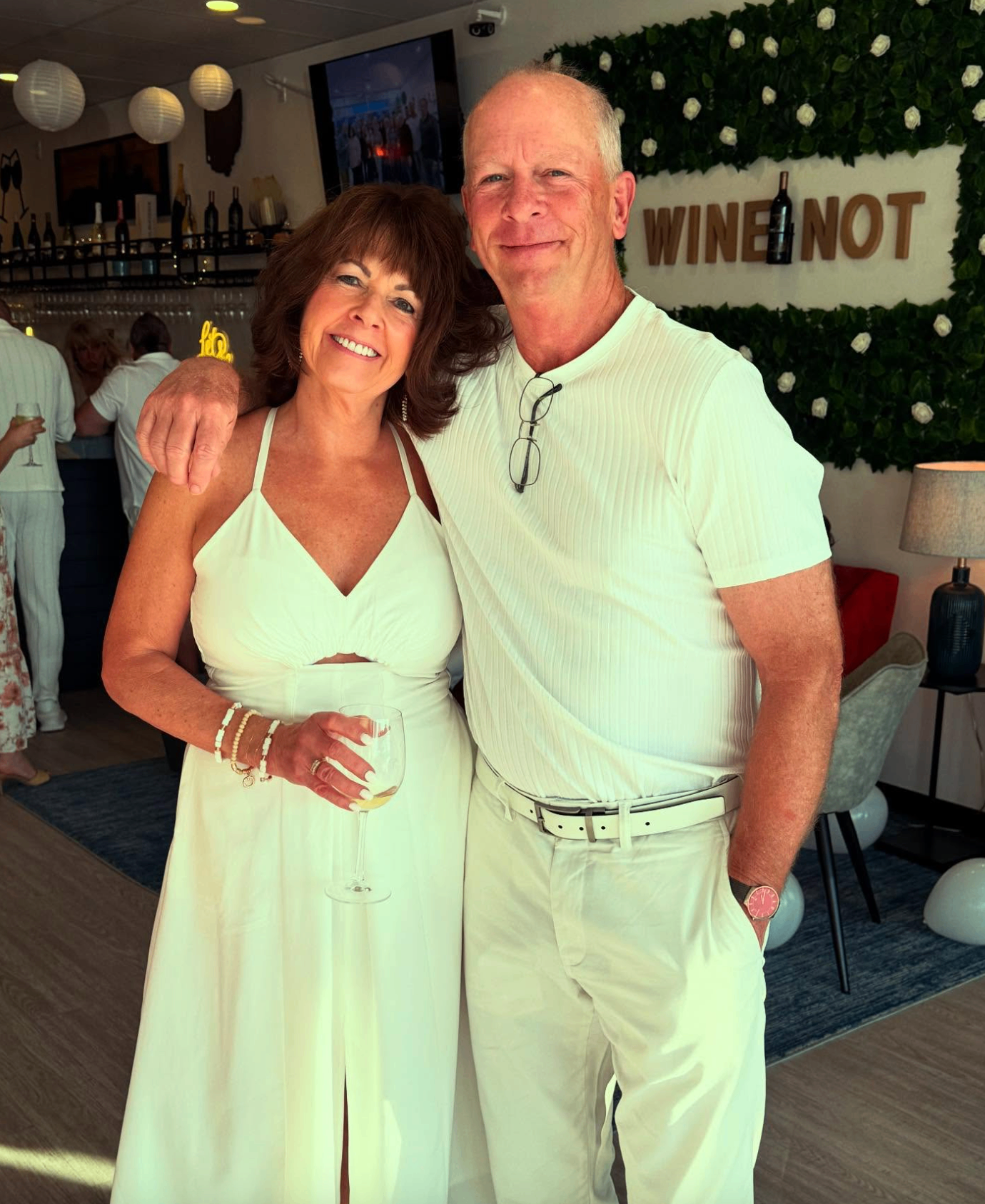 Couple in white posing in front of the Wine Not flower wall